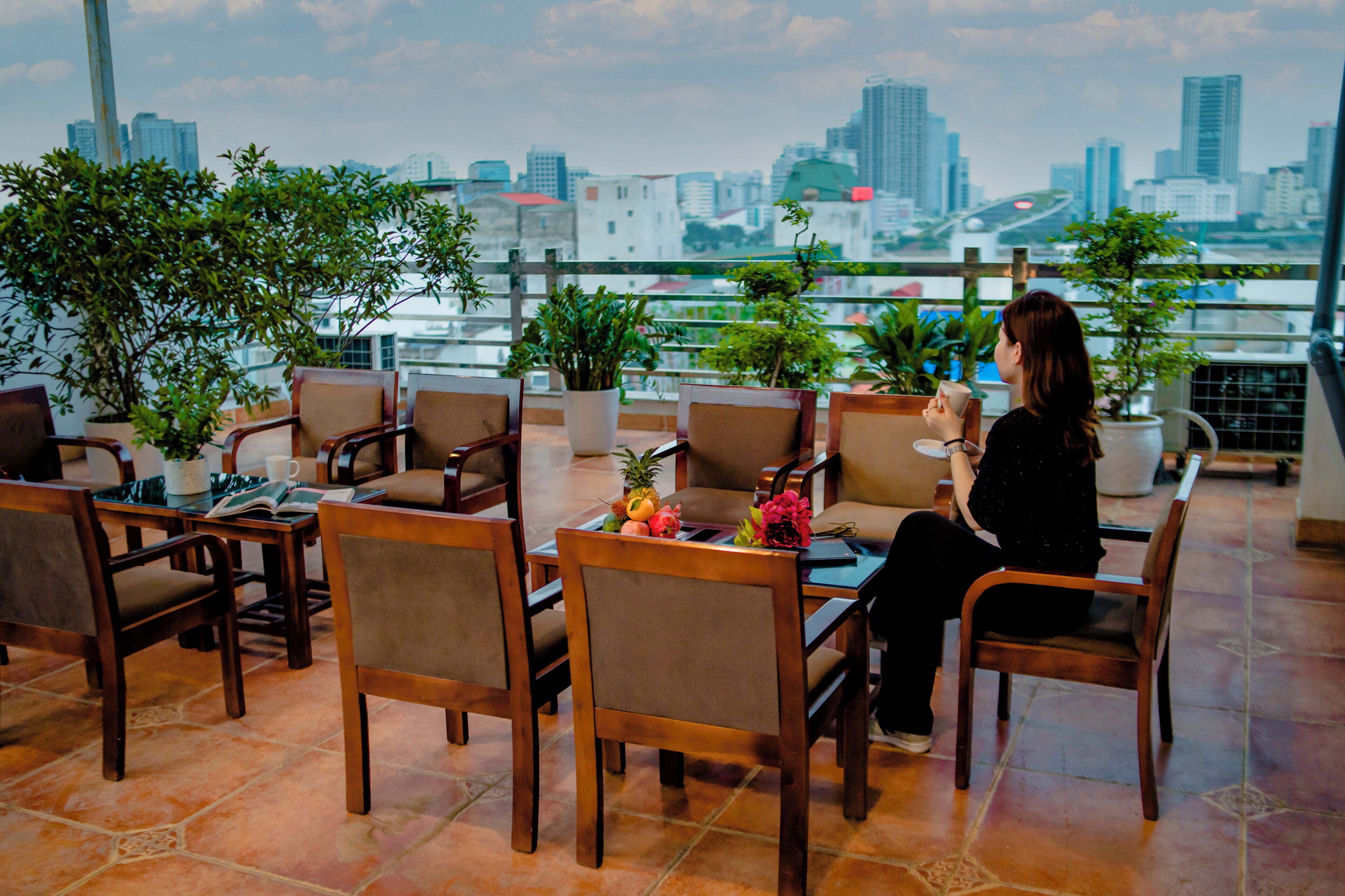 A rooftop seating area with wooden chairs and tables, potted plants, and a view of a city skyline.