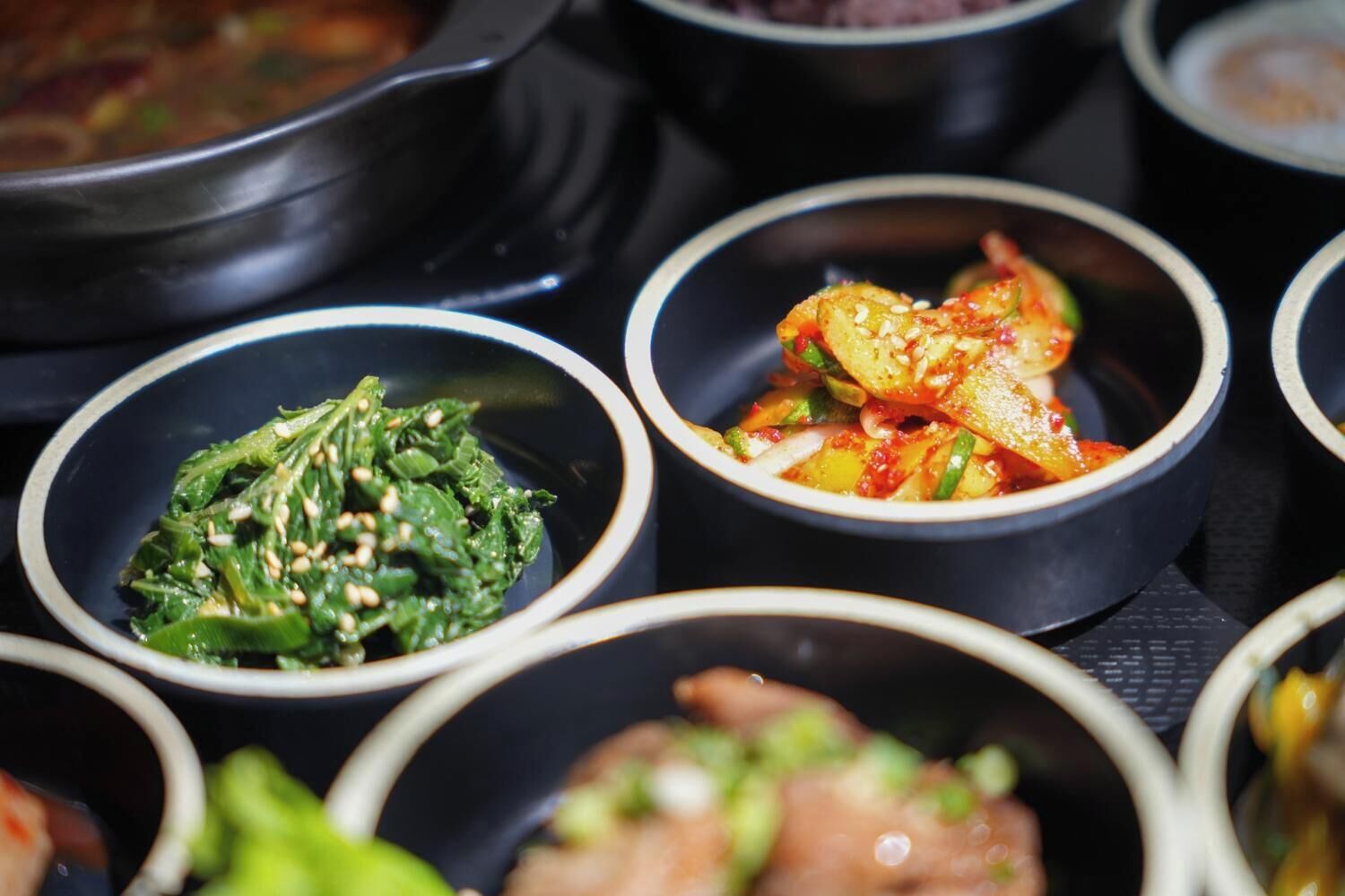 Diversidad de platos coreanos servidos en cuencos negros pequeños, incluyendo kimchi y un salteado picante.