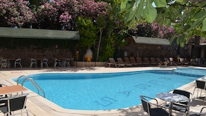 A swimming pool with lounge chairs and tables, surrounded by lush greenery and flowering plants. - Megas Hotel (Ayvalik)