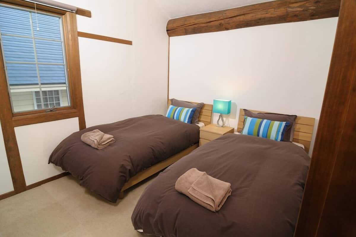 Een slaapkamer met twee eenpersoonsbedden, houten hoofdborden, een nachtkastje met een lamp en een raam met zonwering.