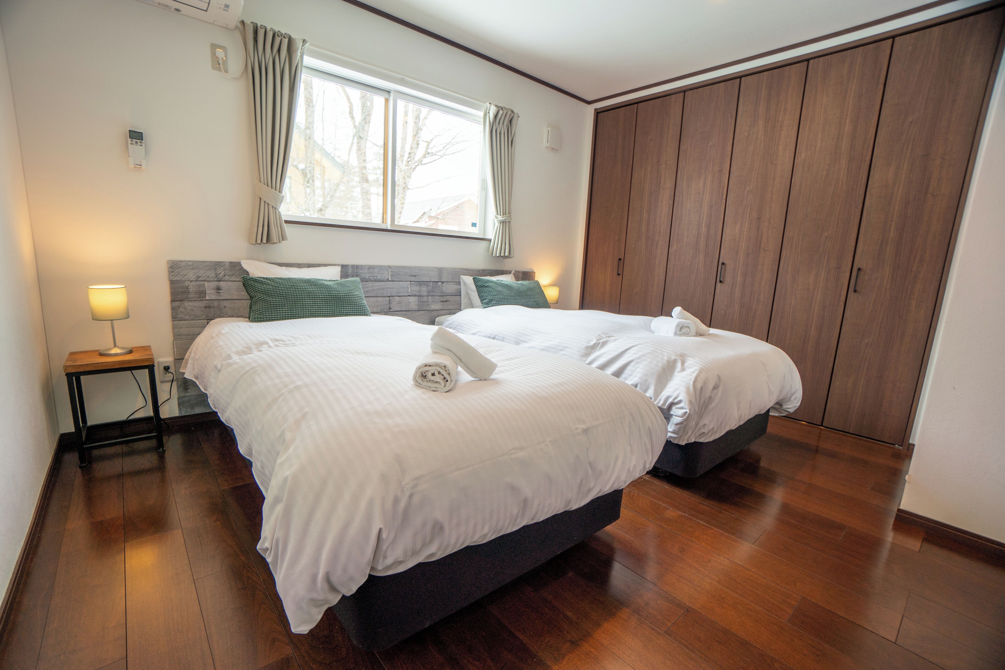 A bedroom with two beds, a wooden floor, a window with curtains, and a wardrobe.