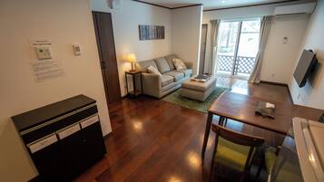 A modern living room with a sofa, a coffee table, a dining table, and a TV. There is a wooden cabinet and a lamp.