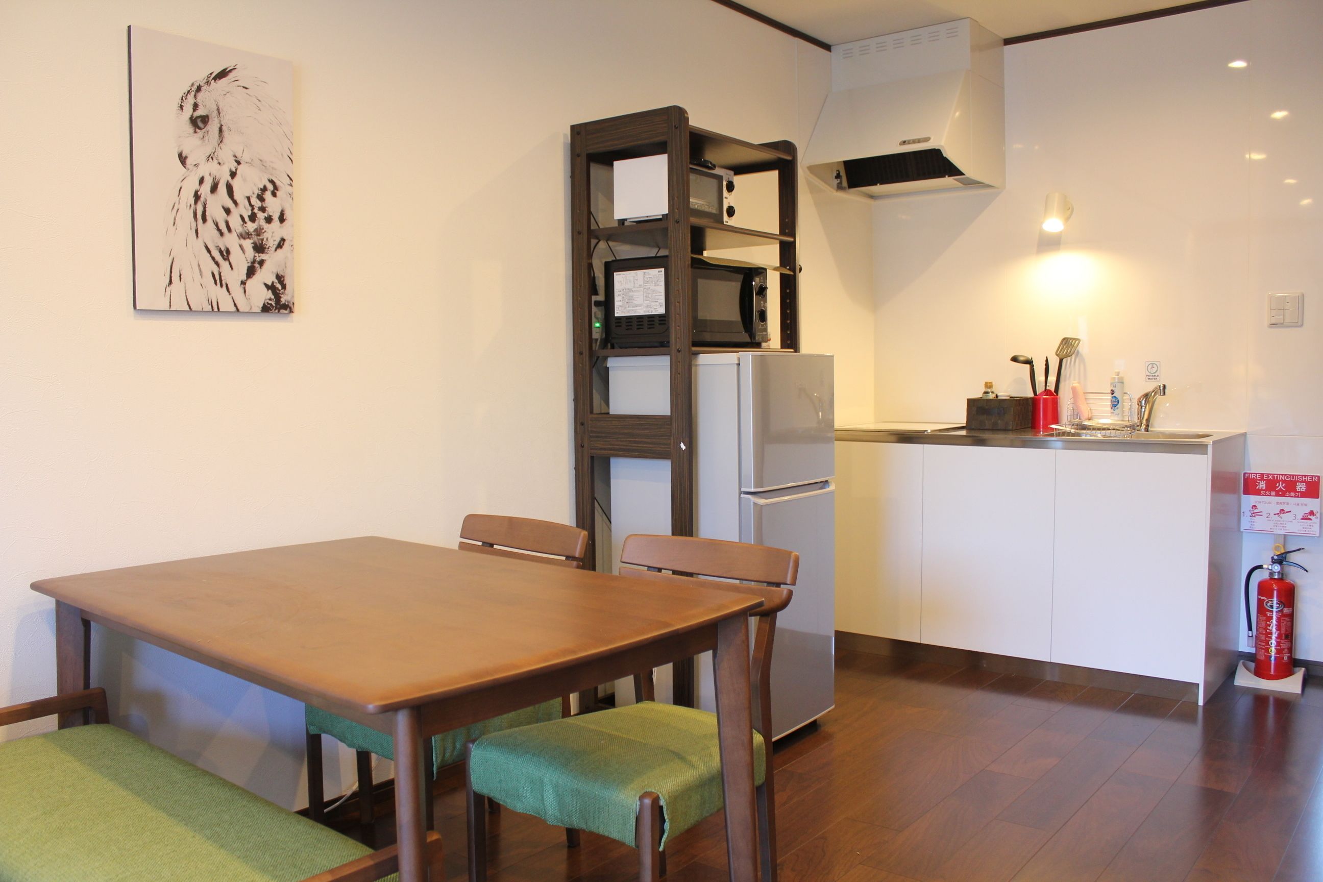 Una cocina compacta con microondas, refrigerador y lavamanos. Hay una mesa de comedor con sillas y un cojín verde. Un extintor está montado en la pared.