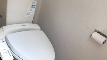 A white toilet with a control panel, a toilet paper holder on the wall, and a small trash bin.