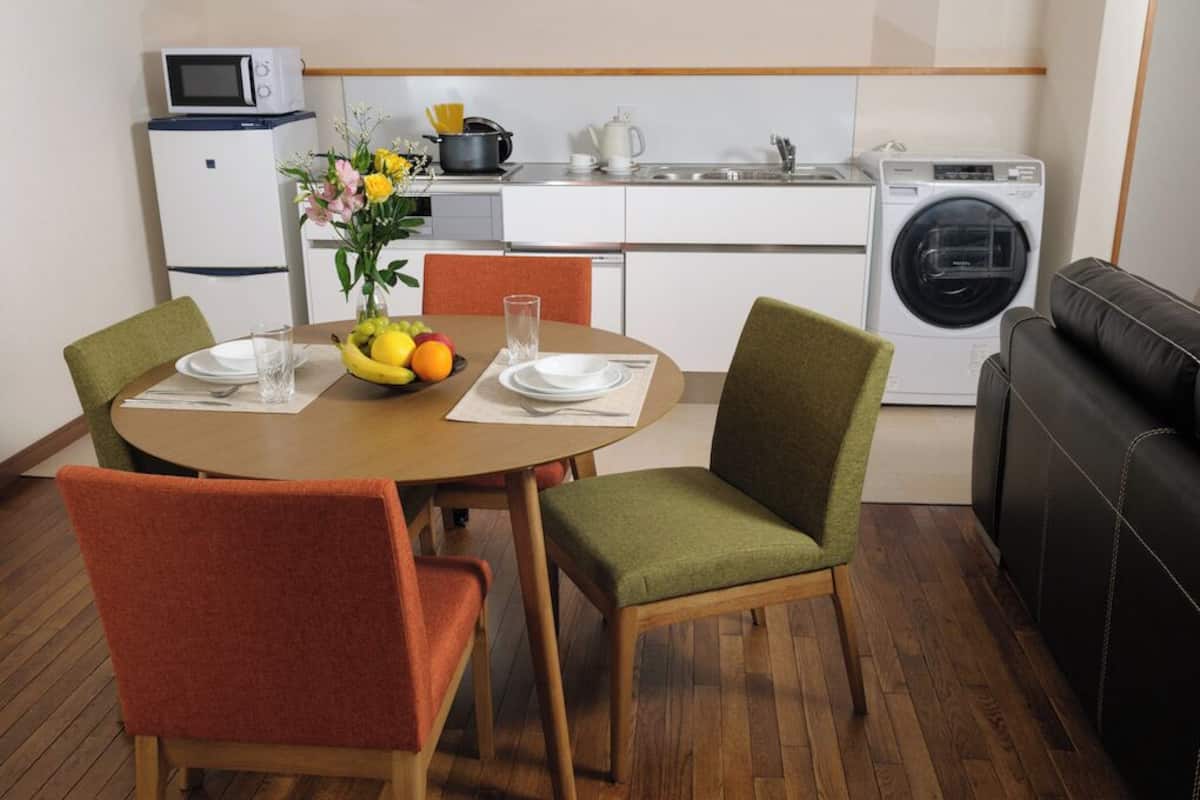 Een functionele keuken met een magnetron, kookplaat, spoelbak en wasmachine. Er is een eettafel met stoelen, een vaas met bloemen en een schaal met fruit.