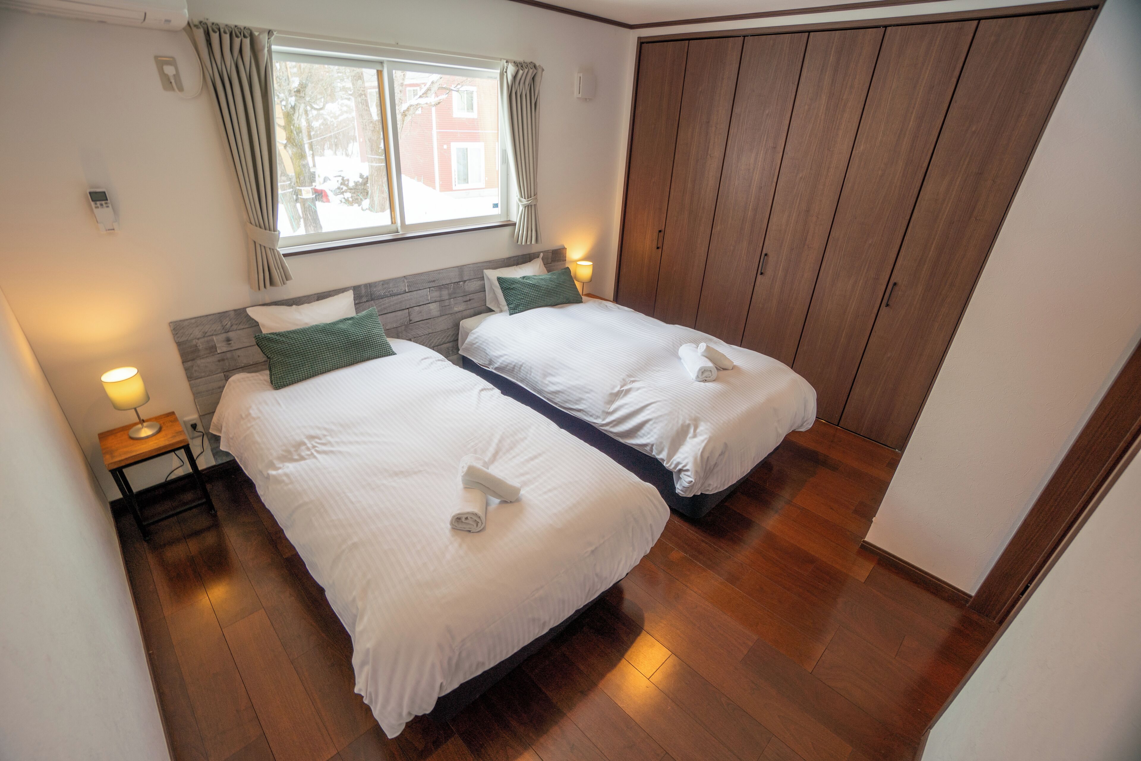 A hotel room with two beds, wooden flooring, a window with curtains, and a wardrobe.