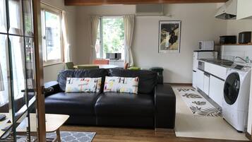A compact living space with a black leather sofa, a dining table, a kitchen area with a washing machine, and a large window providing natural light.