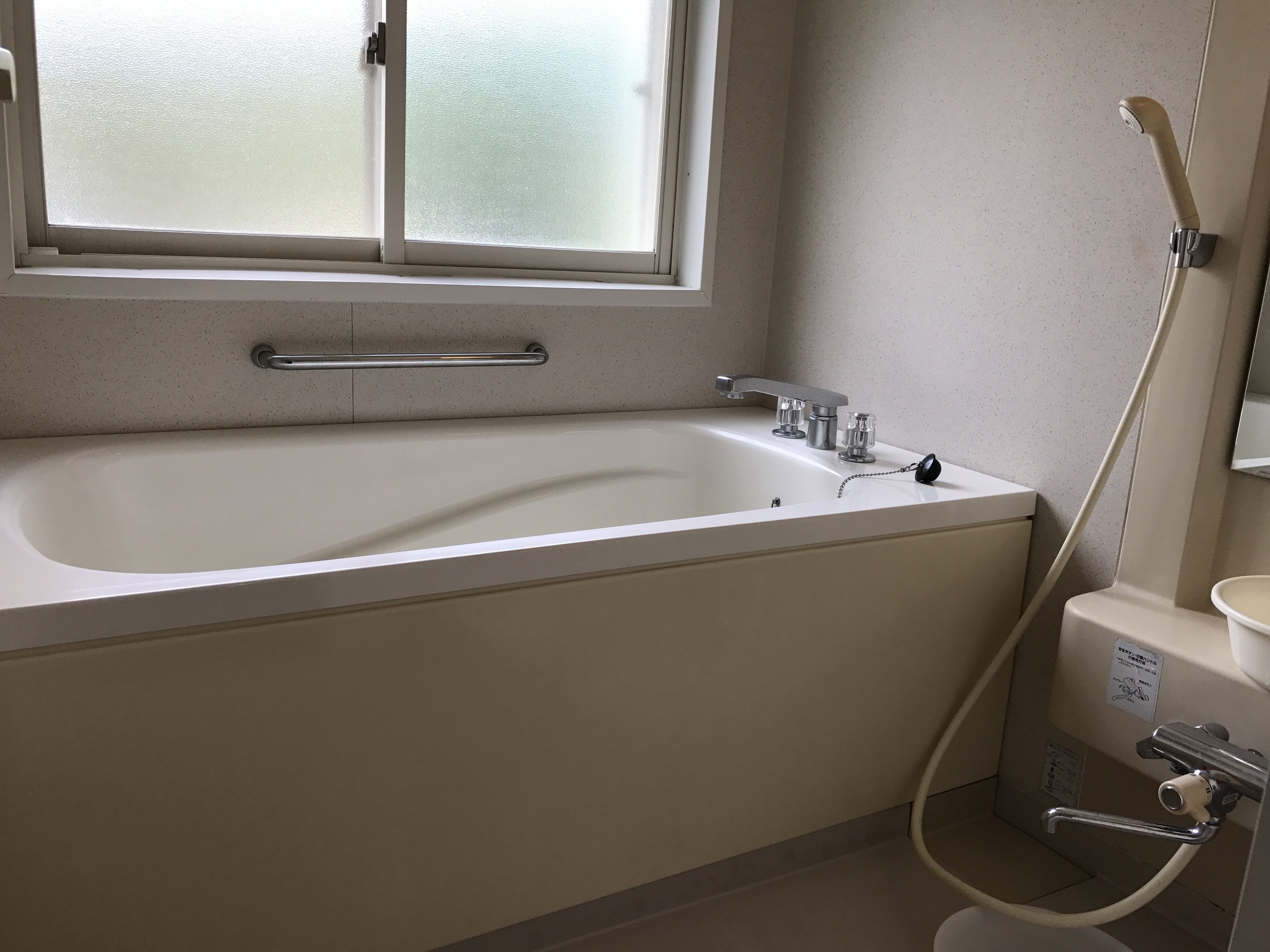 A bathroom with a bathtub, a window, a handrail, a faucet, and a shower hose.