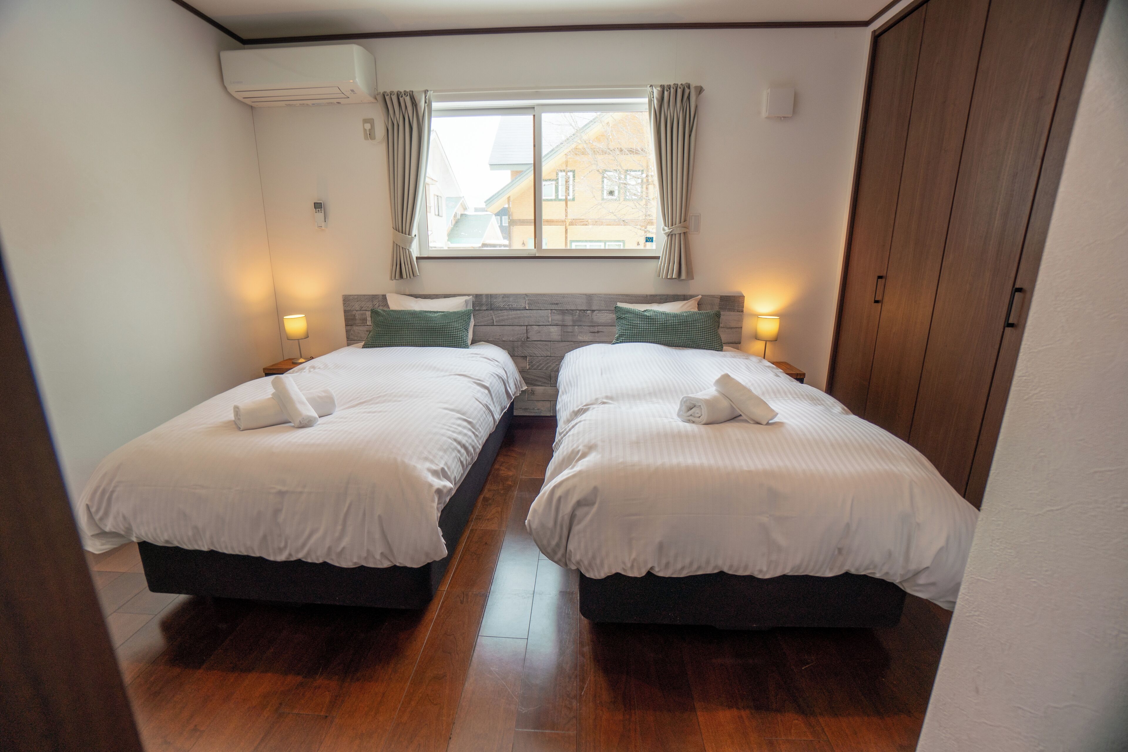 Ett hotellrum med två sängar, trägolv, ett fönster med gardiner och sänglampor.