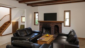 A living room with a fireplace, black leather sofas, a wooden staircase, and a flat-screen TV mounted above the fireplace.