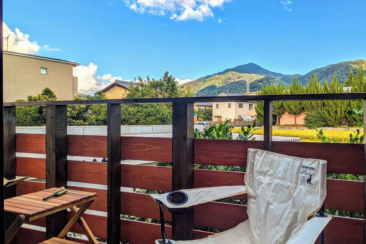 A balcony with a wooden fence, a table, and a chair. In the background, there are buildings and mountains.