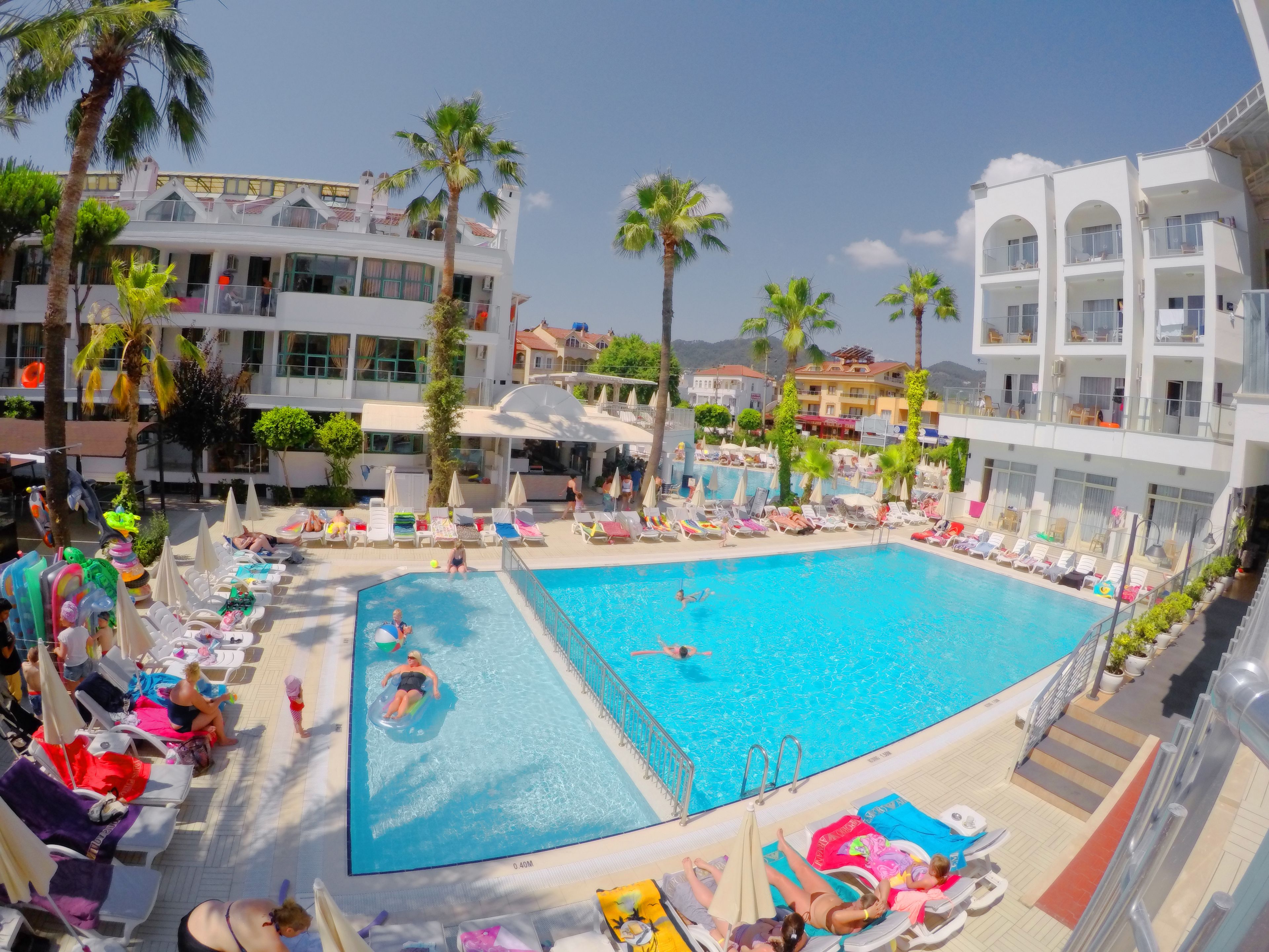 A large swimming pool surrounded by lounge chairs and umbrellas, with palm trees and a multi-story building in the background.