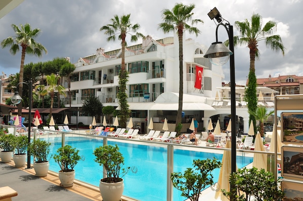 A poolside area with lounge chairs and umbrellas, surrounded by palm trees and a large building with a flag. - Club Atrium Marmaris (Marmaris)