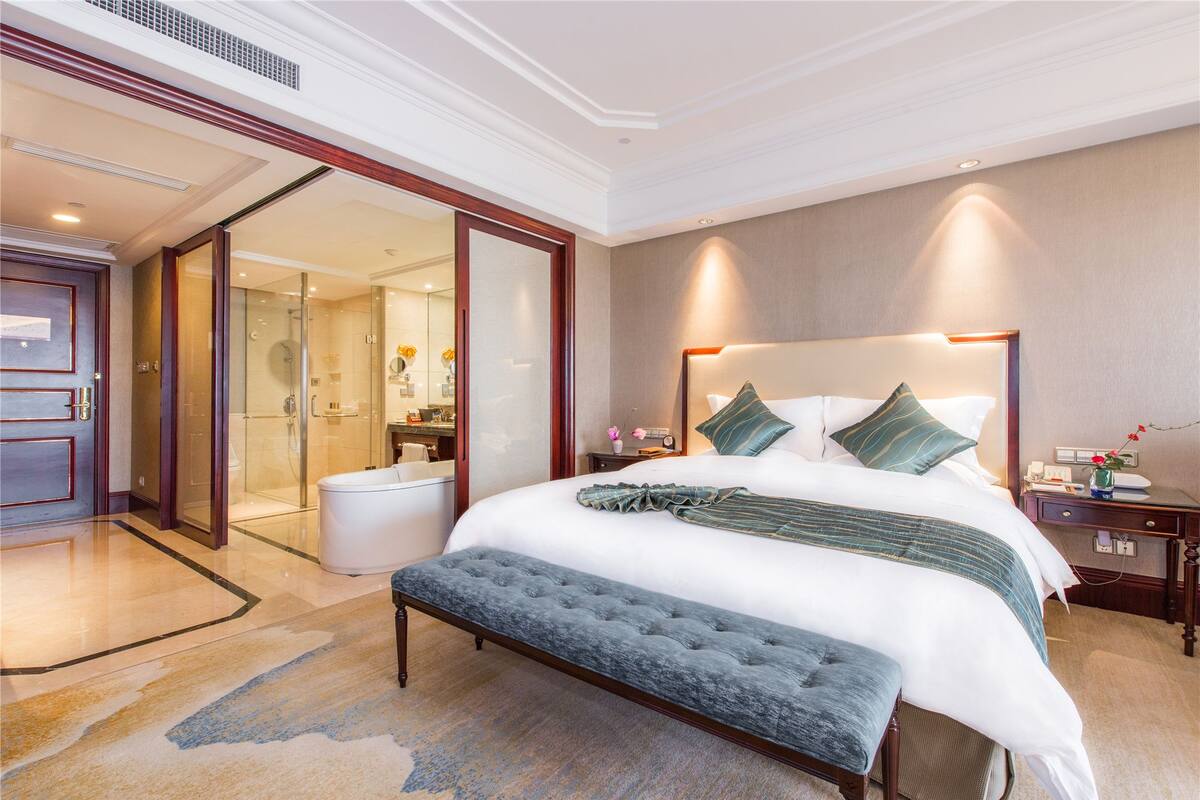 A hotel room with a large bed, a bench, a nightstand, and a bathroom visible through an open door.
