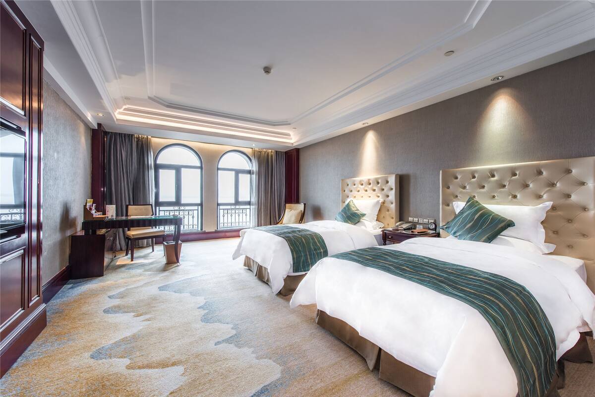 A hotel room with two beds, a desk, and a view of the city through large windows.