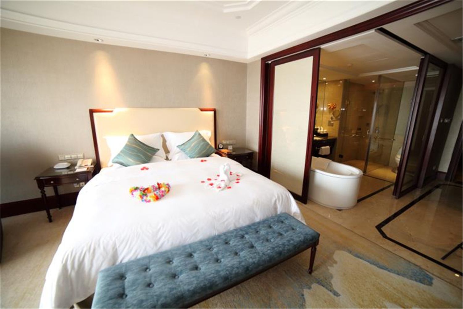 A hotel room with a large bed, a bedside table, a bench, a glass-enclosed bathroom, and a door leading to another room.