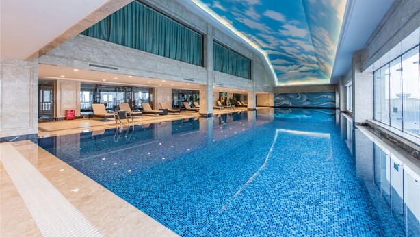 A large indoor swimming pool with clear blue water, surrounded by lounge chairs and a high ceiling with a sky mural. - Qianjiang Junting Hotel, Haining (Jiaxing)