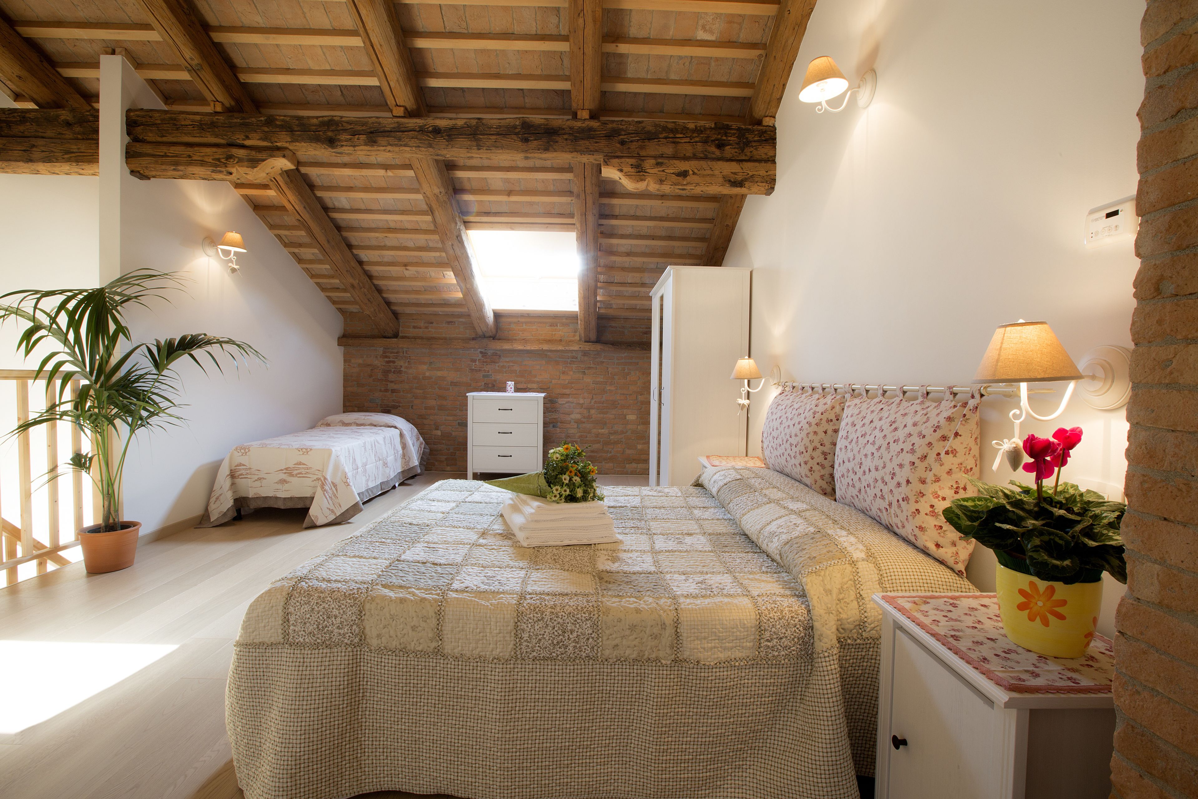 A bedroom with a large bed, a bedside table with a lamp, a potted plant, and a brick wall.