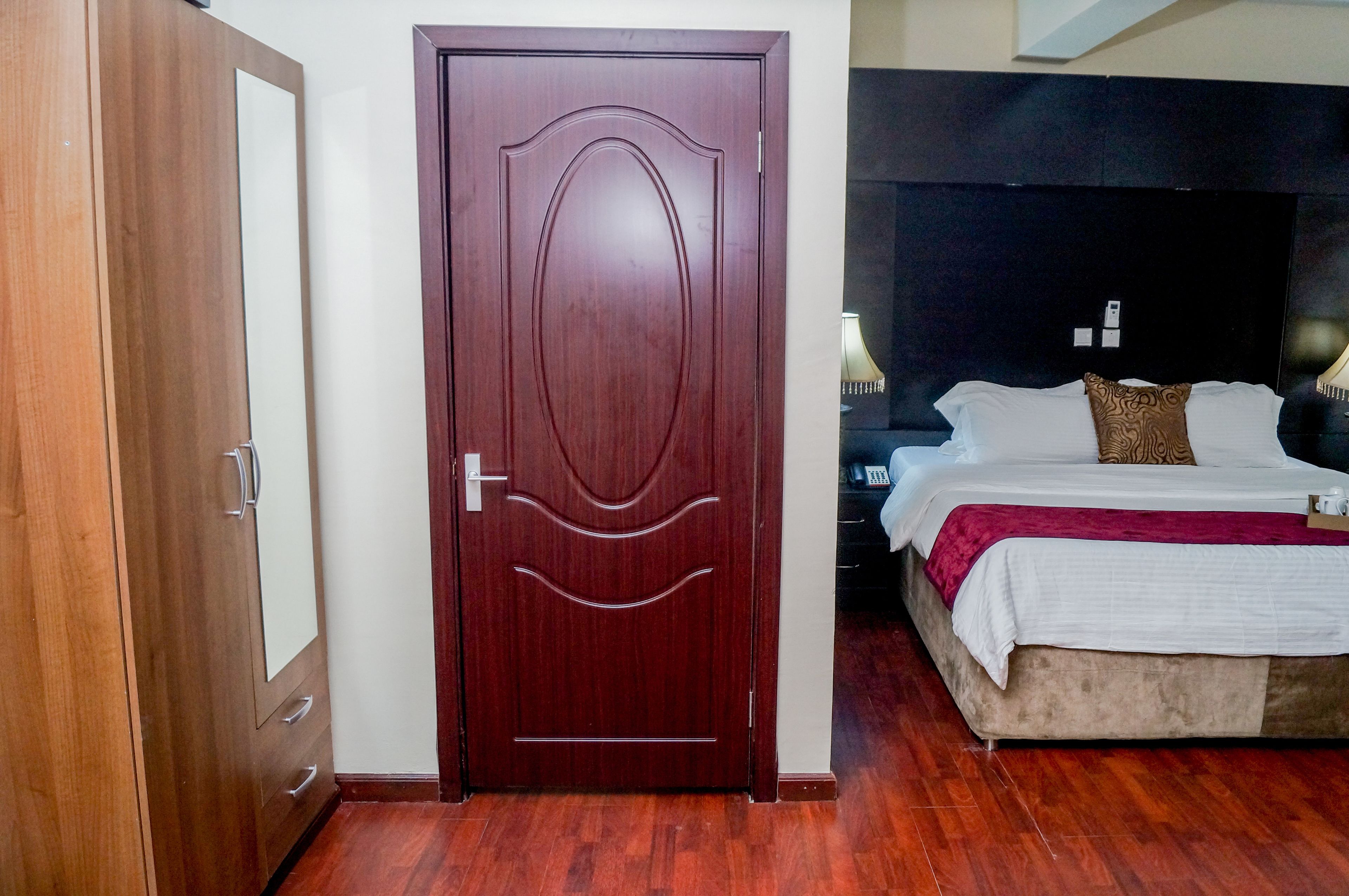 A hotel room with a wooden door, a bed with white bedding and a red throw, and a nightstand with a lamp.