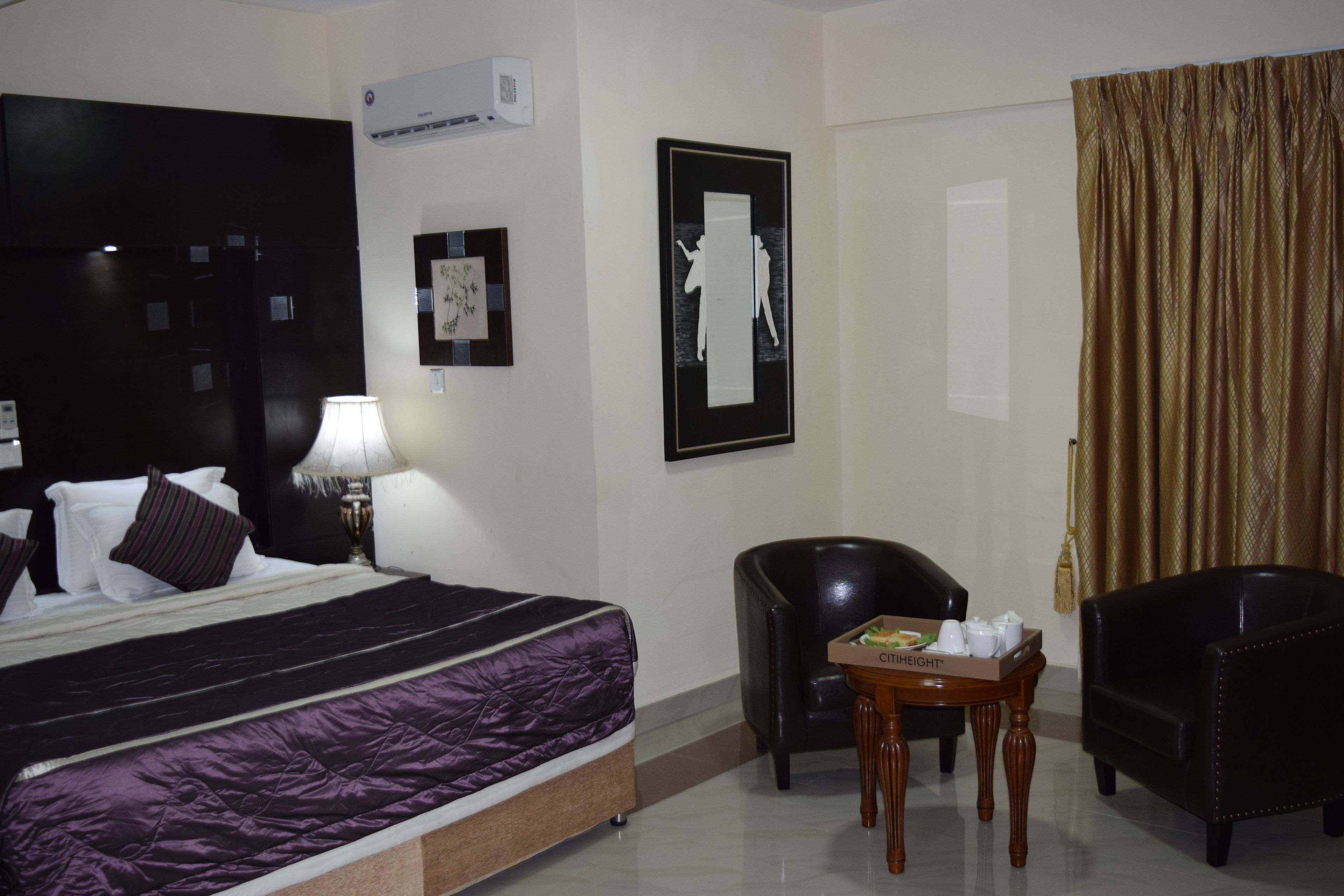 Una habitación de hotel con una cama, un escritorio con silla, una lámpara, un cuadro enmarco y una ventana con cortinas.