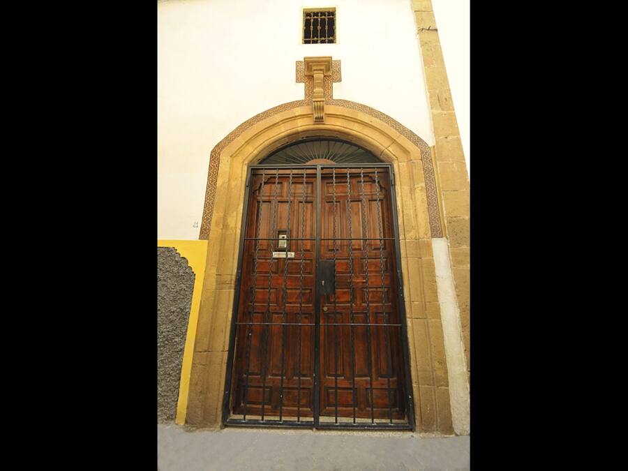 Une grande porte en bois richement décorée, surmontée d’un portail en métal, le tout intégré dans une arche en pierre.