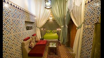 A narrow room with a patterned tile wall, a green sofa with red cushions, a small table with a tray, and a hanging lantern.