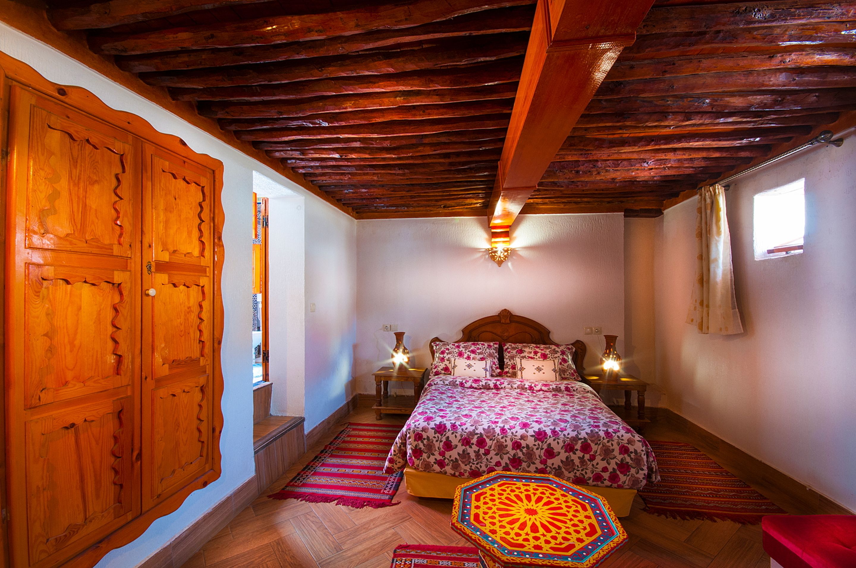 A bedroom with a wooden ceiling, a large bed with floral bedding, two bedside lamps, and a red chair.