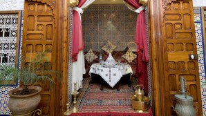 A traditional Moroccan interior with a wooden door, red and white drapes, a patterned rug, and a small table with a lantern. - Riad Dar Jabador (Salé)