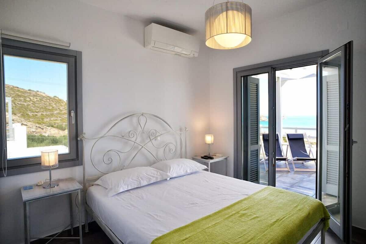 A bedroom with a bed, a nightstand, a lamp, and a view of the sea through an open door.