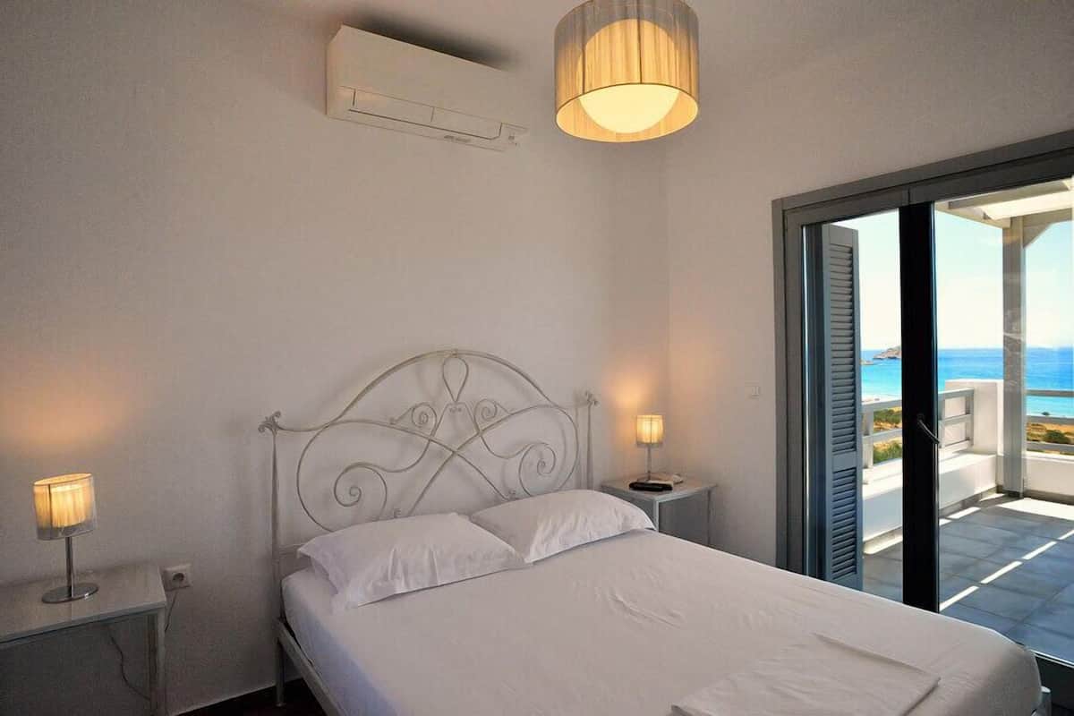 A bedroom with a bed, bedside lamps, a ceiling air conditioner, and a view of the sea through an open door.