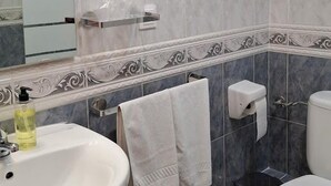 Une salle de bain avec un lavabo, des toilettes et une douche. Une serviette est accrochée au mur et un distributeur de savon est posé sur le lavabo.