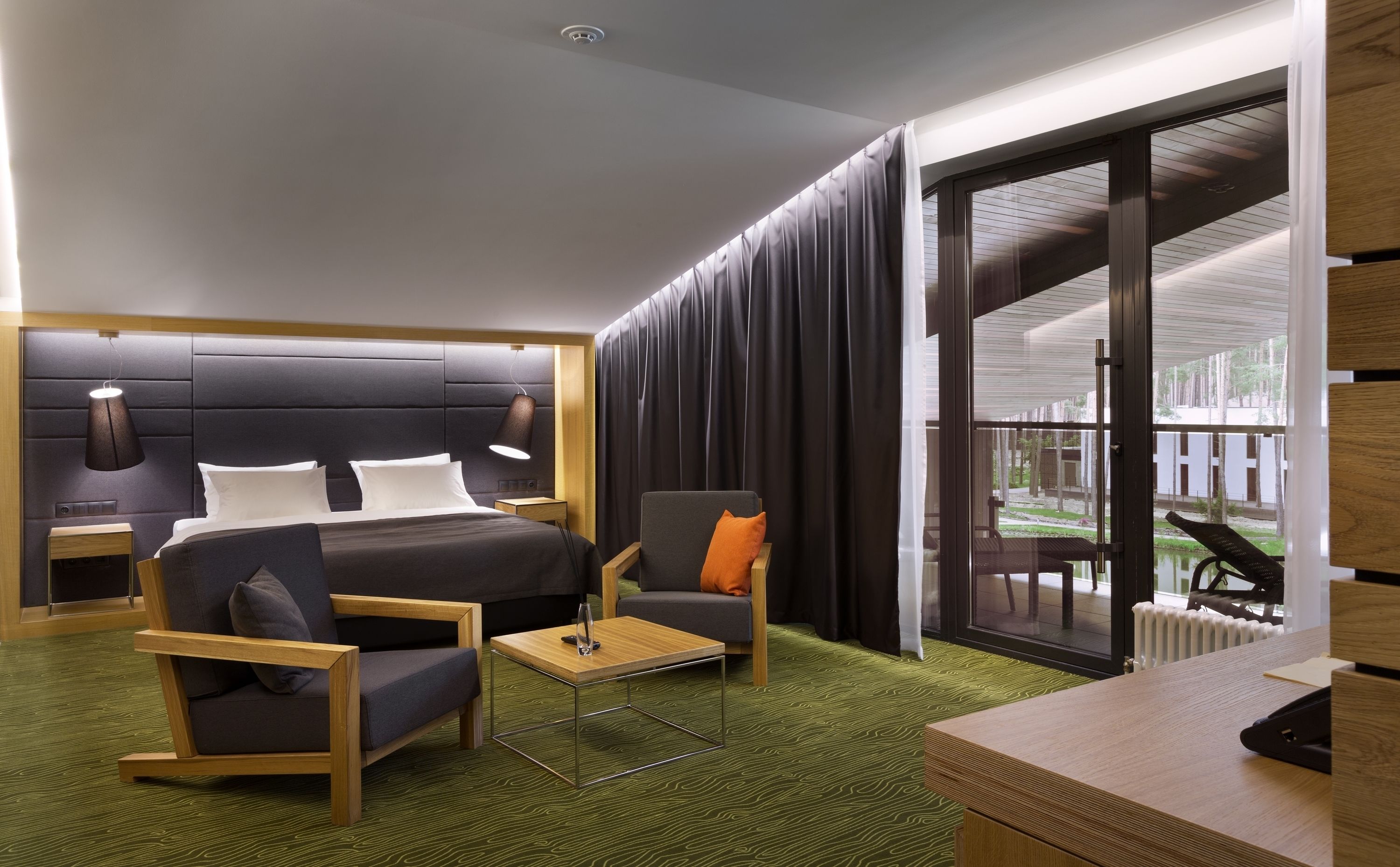 A modern hotel room with a large bed, a desk, a chair, and a view of the outdoors through a sliding glass door.