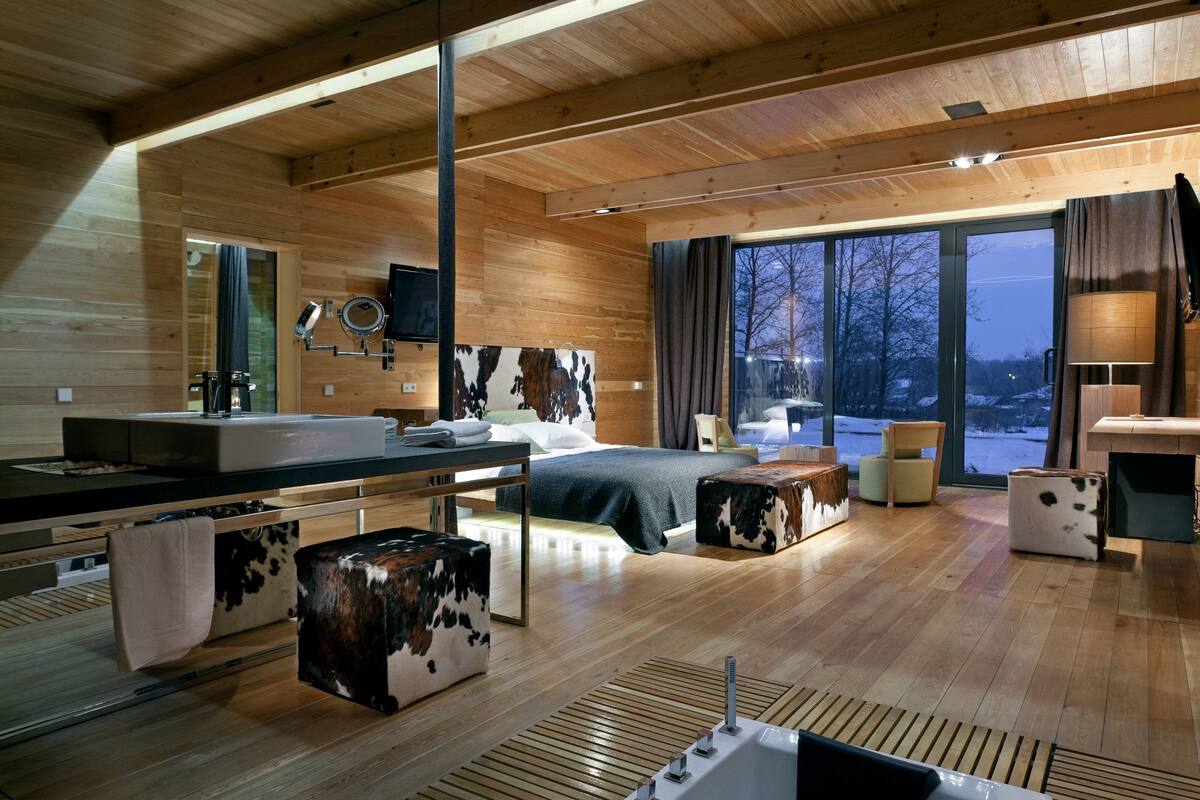 A modern wooden interior with a large bed, a bathroom with a sink and mirror, and a view of the outdoors.