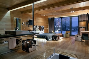 Interior moderno em madeira com uma cama grande, banheiro com pia e espelho, e vista para o exterior.