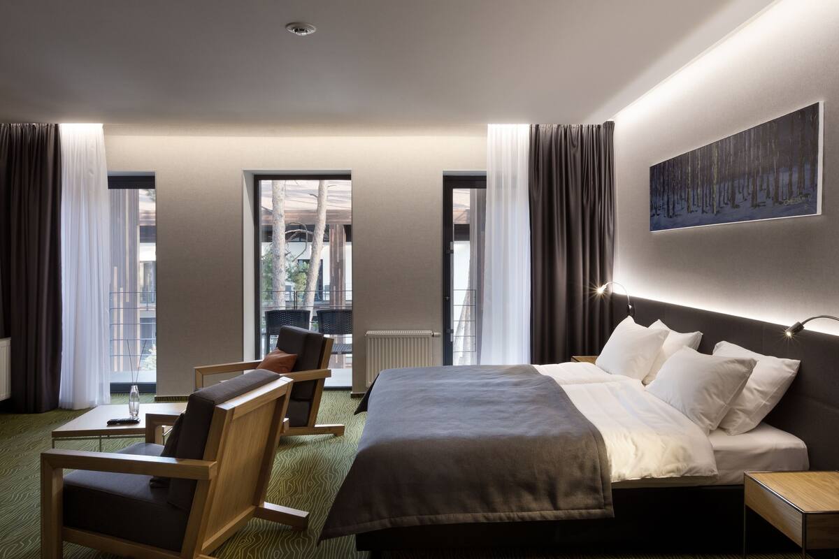 A modern hotel room with a large bed, a desk, and a chair. There are windows with curtains, a painting on the wall, and a view of the outside.