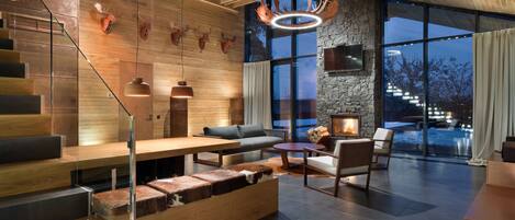 A modern interior with a wooden staircase, a stone fireplace, and a large window offering a view of the outdoors.