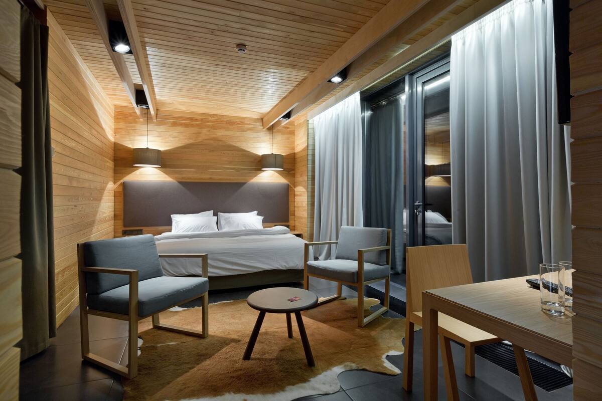 A modern hotel room with a wooden ceiling, a large bed with white linens, two chairs, a small round table, and a glass of water.