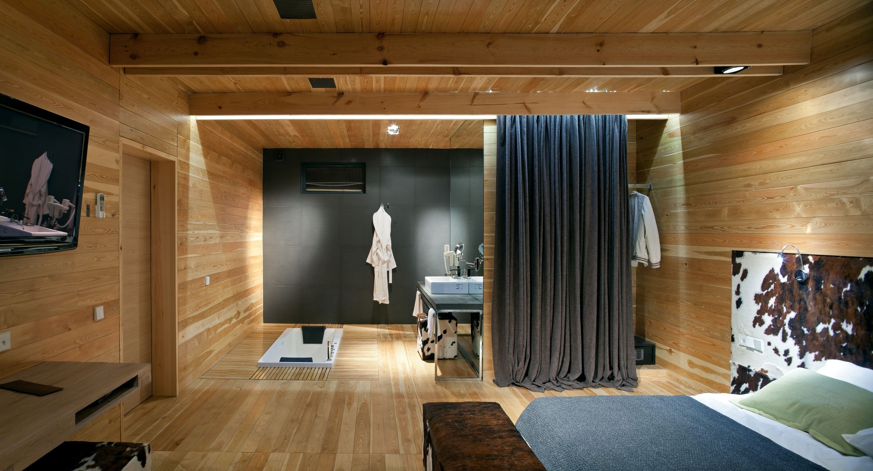 A modern wooden interior with a bed, a small sitting area, a bathroom with a mirror and towel rack, and a television on the wall.
