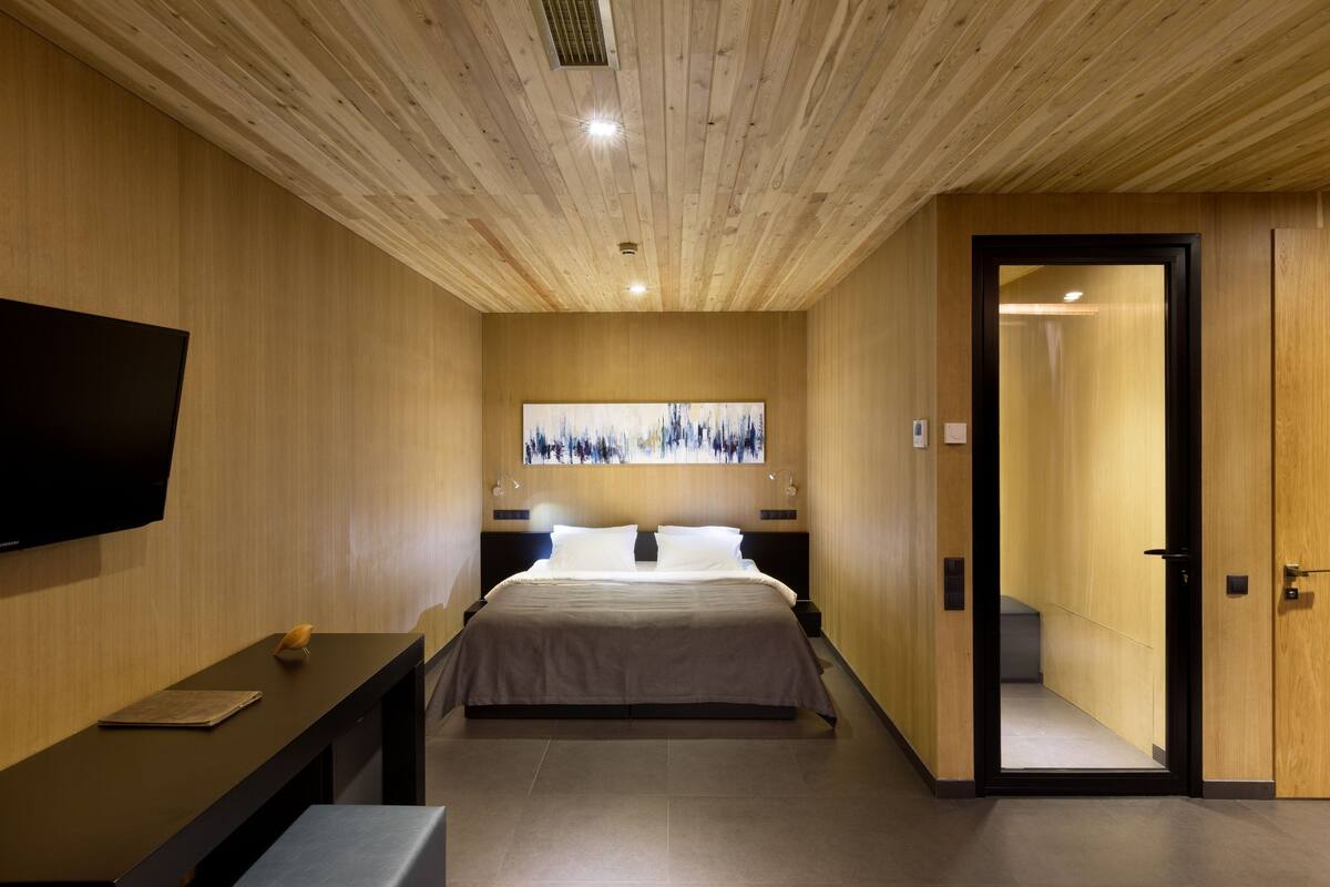 A modern hotel room with a wooden ceiling, a large bed, a flat-screen TV, and a painting on the wall.
