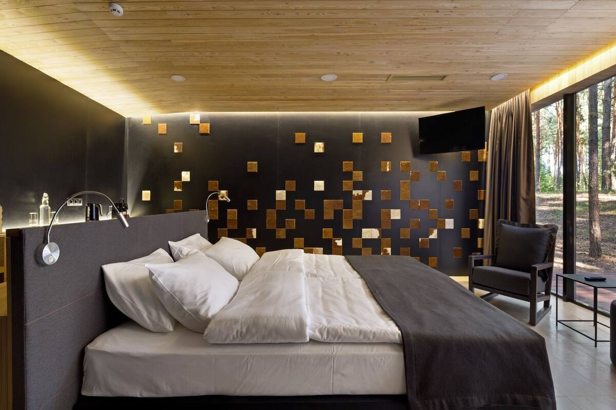 A modern bedroom with a large bed, a bedside table, a chair, and a view of trees through large windows.