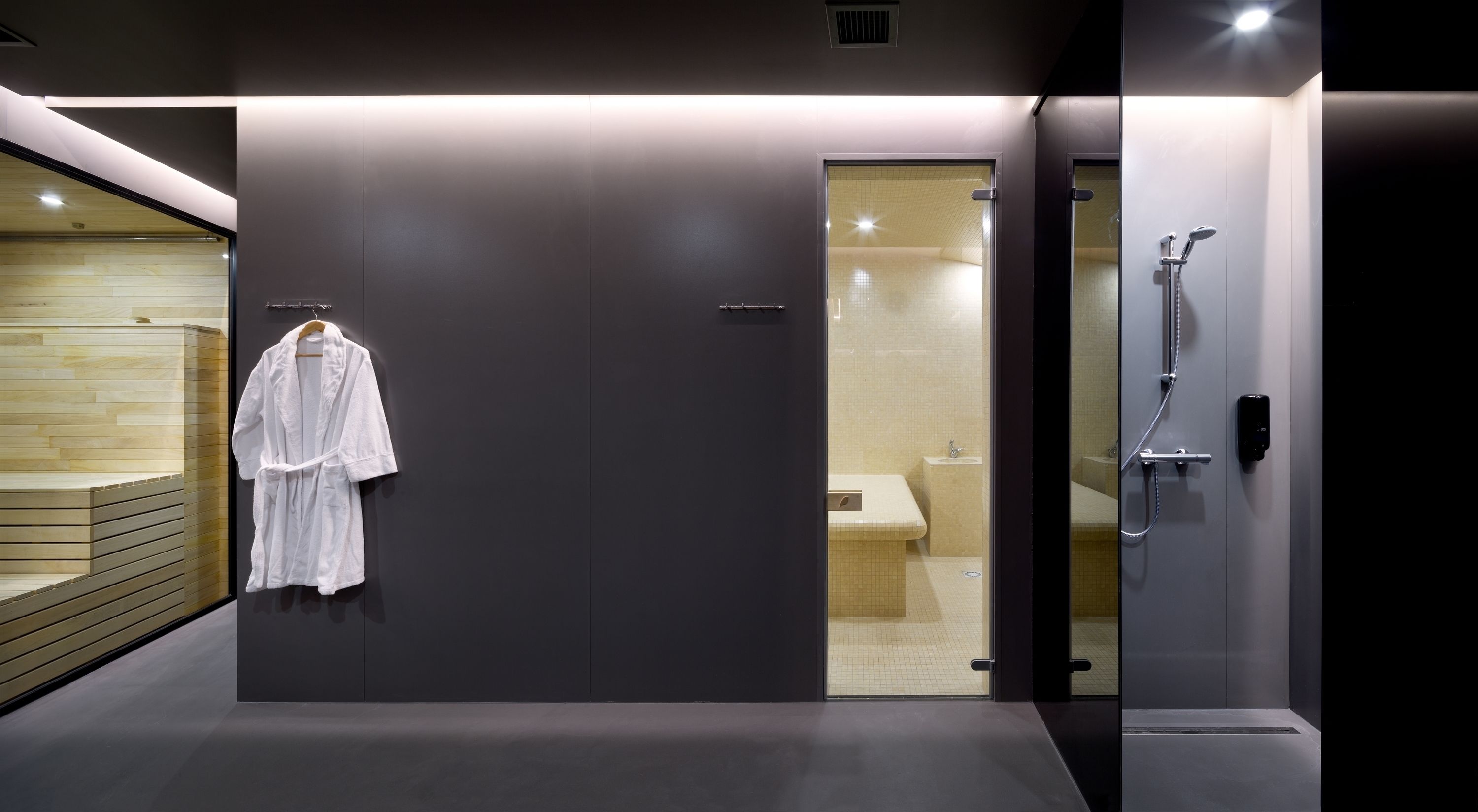 A modern bathroom with a glass shower enclosure, a wooden bench, and a white robe hanging on the wall.