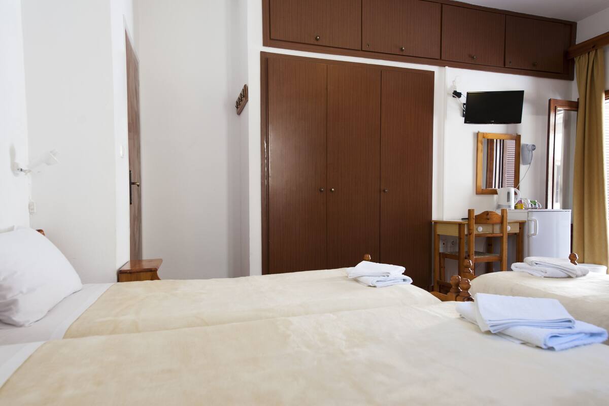 A hotel room with two beds, a wooden wardrobe, a desk with a chair, a television, and a mirror.