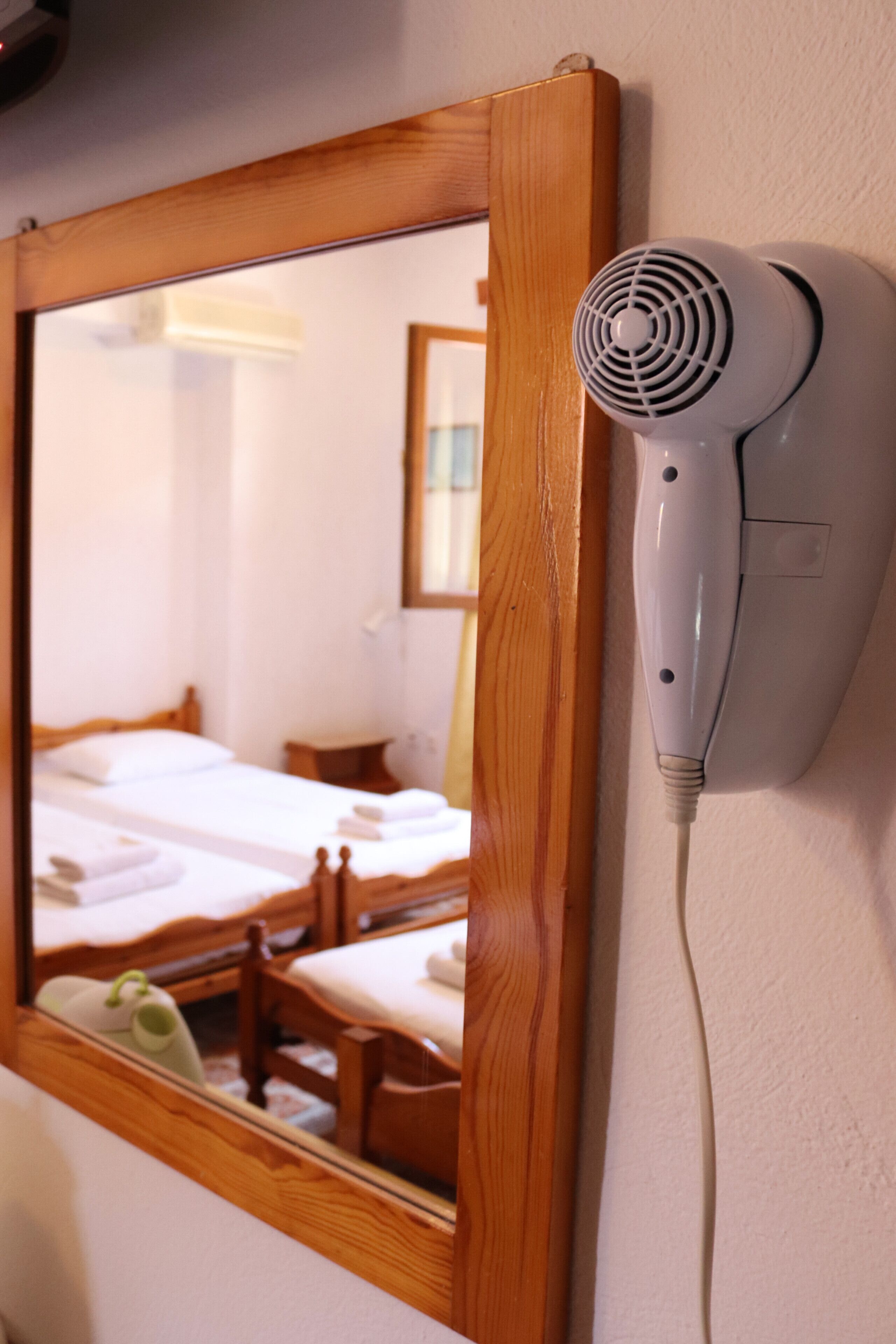 A hair dryer mounted on a wall next to a wooden-framed mirror reflecting a room with a bed and a chair.