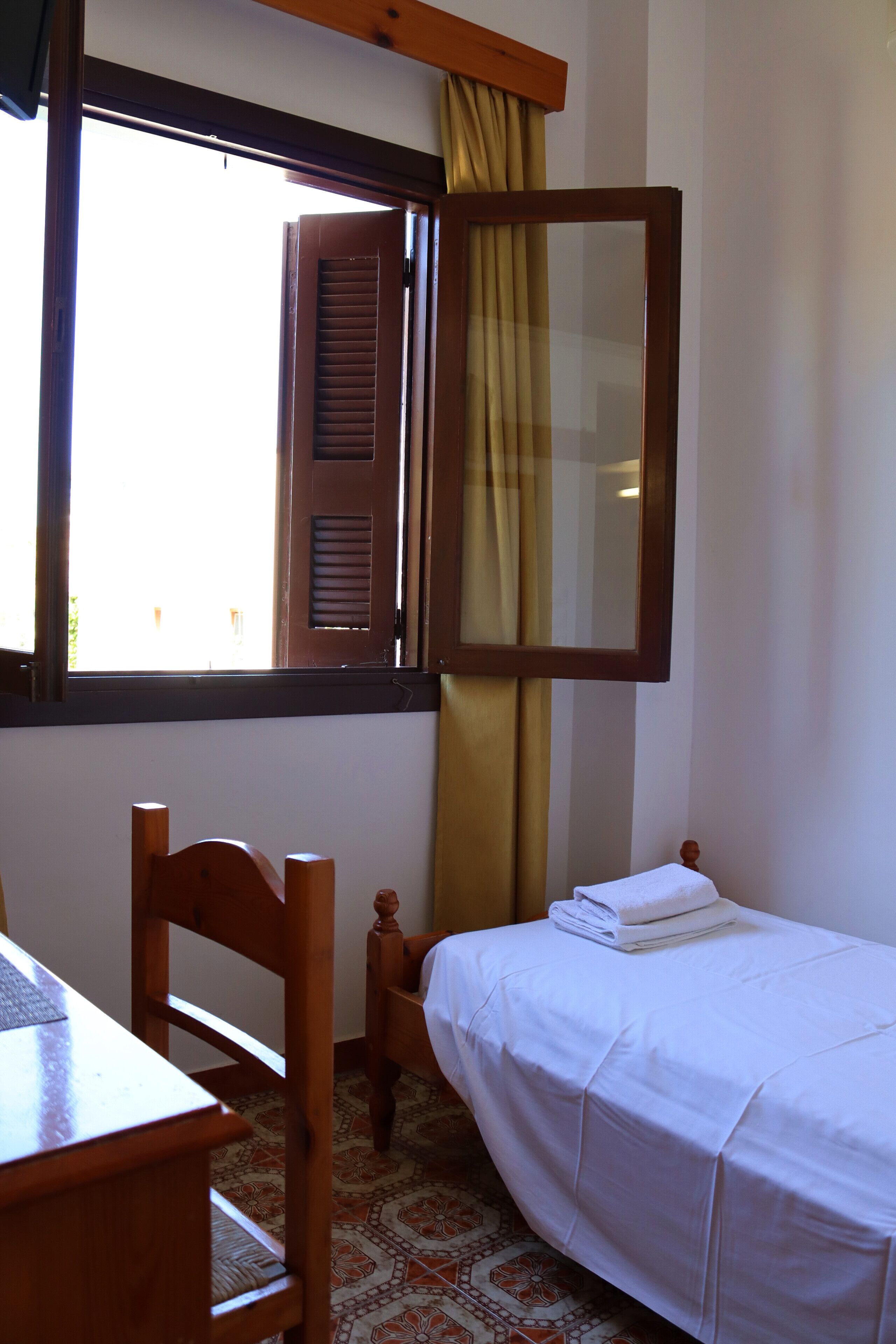 Um quarto pequeno com uma cama, uma cadeira, uma mesa e uma janela com cortinas.