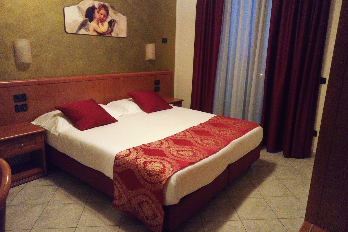 Une chambre d’hôtel avec un grand lit, deux oreillers rouges, une tête de lit en bois, une couvre-lit rouge et or, une table de chevet, une lampe fixée au mur et une fenêtre avec des rideaux.