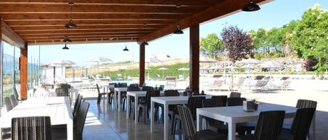 An outdoor dining area with tables and chairs, a covered roof, and a view of a pool and garden.