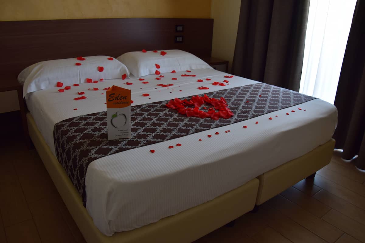 Un lit bien fait, décoré de pétales de rose et agrémenté d’une carte, une couvre-lit à motifs et une tête de lit en bois.
