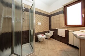 A bathroom with a glass shower enclosure, two toilets, a bidet, and a sink with a window above. - Hotel Residence Eden (Mozzagrogna)