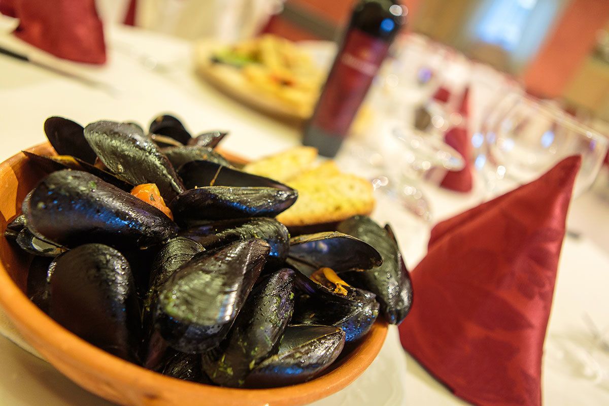 A bowl of mussels on a table with a red napkin and a bottle in the background.