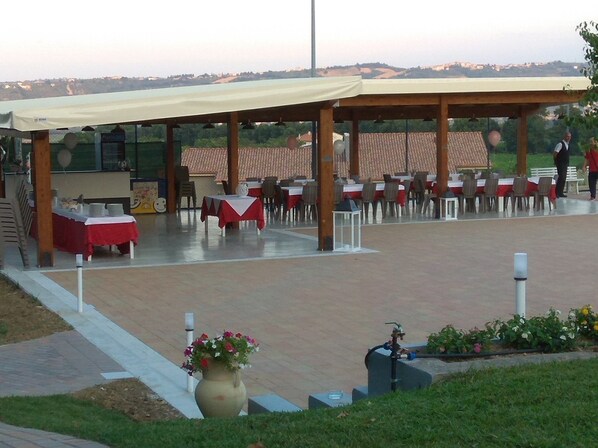 An outdoor dining area with tables and chairs under a covered pavilion, a large vase with flowers, and a water fountain.